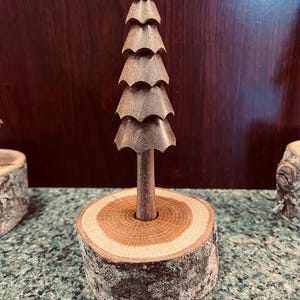 May include: A handcrafted wooden Christmas tree sculpture. The tree is brown and has a layered design, mounted on a natural wood base. The base is a cross-section of a tree trunk, with visible growth rings and bark.