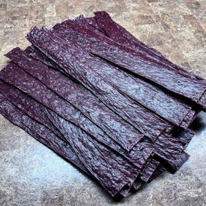 May include: A stack of thin, dark purple, dried fruit leather strips.