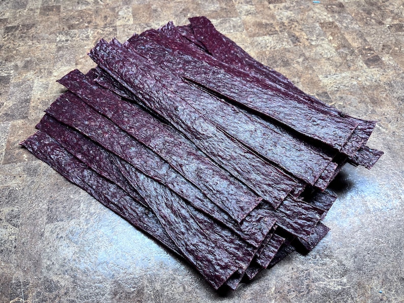 May include: A stack of thin, dark purple, dried fruit leather strips.