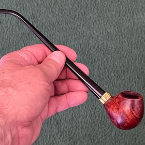 May include: A classic tobacco pipe with a dark, curved stem and a reddish-brown bowl. The bowl has a rich, wood-grain appearance, and a gold-colored band accents the connection between the stem and bowl. The pipe is held in a hand against a green background.