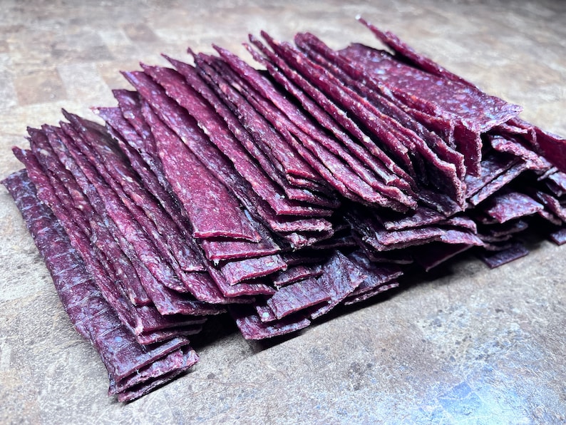 May include: A stack of thin, purple-colored beef jerky strips. The jerky is arranged in a neat pile, with the strips overlapping each other.
