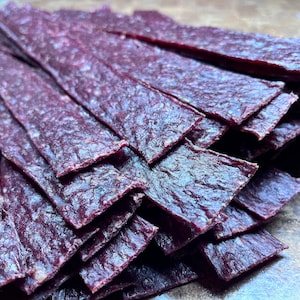 May include: A stack of thin, dark purple beef jerky strips. The jerky is arranged in a neat pile, with the strips overlapping each other.