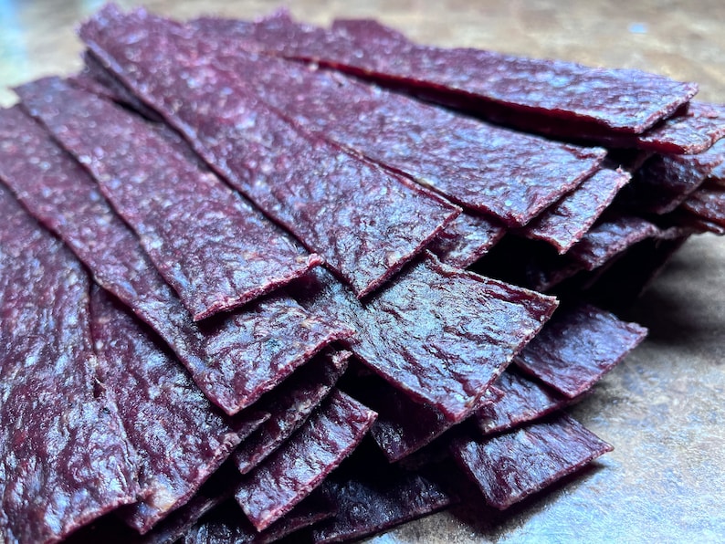 May include: A stack of thin, dark purple beef jerky strips. The jerky is arranged in a neat pile, with the strips overlapping each other.