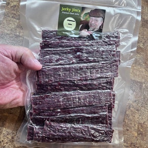 May include: A clear plastic bag filled with thin strips of dark purple beef jerky. The bag has a label that reads "Jerky Jim's Premium Beef Jerky" with a picture of a man wearing a hat and a beard. The label also says "Hickory" and "Established 2023".