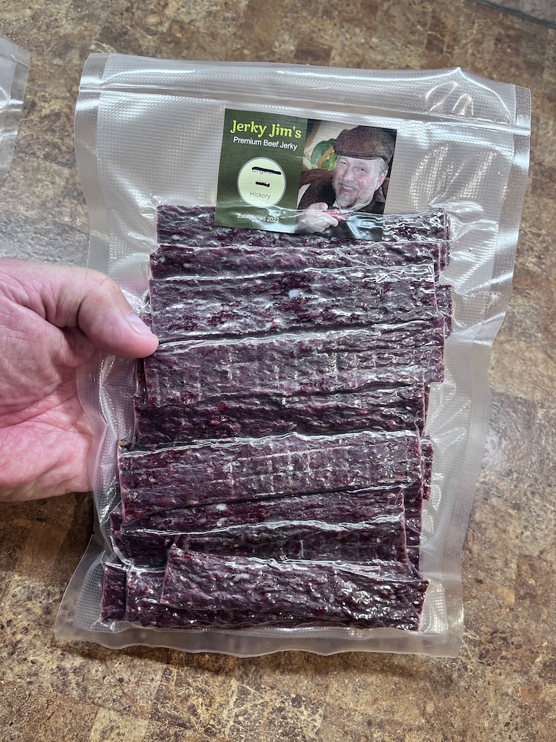May include: A clear plastic bag filled with thin strips of dark purple beef jerky. The bag has a label that reads "Jerky Jim's Premium Beef Jerky" with a picture of a man wearing a hat and a beard. The label also says "Hickory" and "Established 2023".