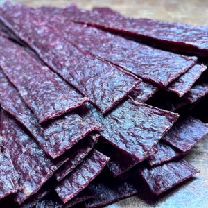 May include: A close-up of a pile of dark red beef jerky strips. The jerky has a textured surface and appears to be stacked in a haphazard manner. The strips are rectangular and vary slightly in size. The overall color is a deep reddish-brown.