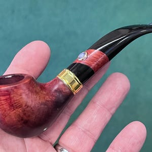 Peut inclure: Une pipe à tabac artisanale avec un foyer brun rougeâtre et un tuyau noir. La pipe présente une bande dorée et un accent décoratif près de l'embout buccal. La pipe est tenue dans une main sur un fond vert.
