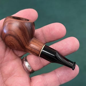 May include: A wooden tobacco pipe with a rounded bowl and a black stem. The pipe is made of a dark, patterned wood and features a gold-colored band where the stem connects to the bowl. The pipe is held in a hand.