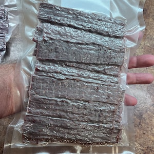 May include: A clear plastic bag filled with a stack of dark brown beef jerky strips.