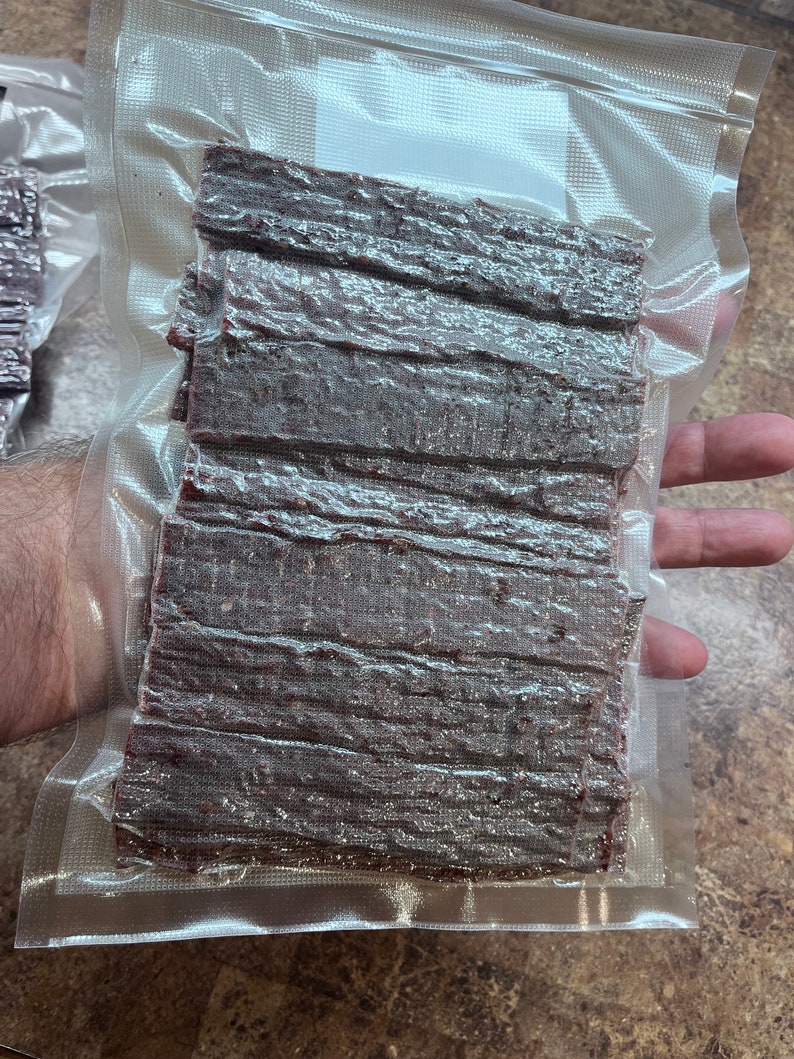 May include: A clear plastic bag filled with a stack of dark brown beef jerky strips.