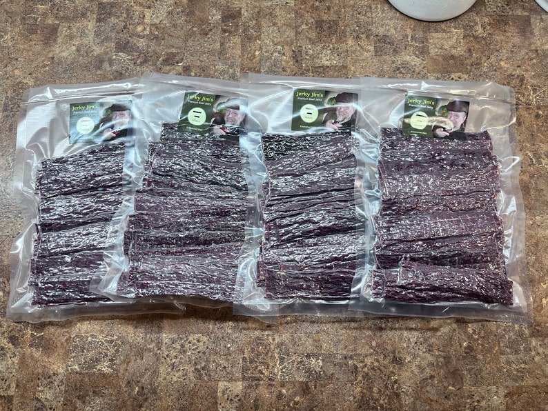 May include: Four clear plastic bags filled with dried meat jerky. Each bag has a label with the text "Jerky Jim's" and a picture of a man.
