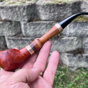 May include: A tobacco pipe featuring a polished reddish-brown wooden bowl with a distinct grain pattern. The stem is composed of alternating segments of light and dark wood, accented by thin silver-colored metal rings, and a black acrylic mouthpiece.