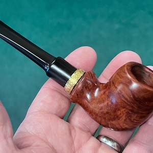 May include: A wooden tobacco pipe with a curved bowl and a black stem. The bowl is a reddish-brown color with a natural wood grain pattern. A gold-colored band with a decorative pattern accents the pipe. The pipe is held in a hand.