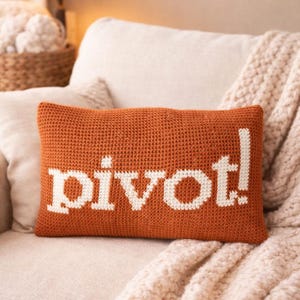 Könnte beinhalten: Ein orangefarbenes Häkelkissen mit dem Wort "pivot!" in weißen Buchstaben. Das rechteckige Kissen liegt auf einem beigen Sofa mit einer cremefarbenen Decke. Eine Lampe und ein Korb befinden sich im Hintergrund.