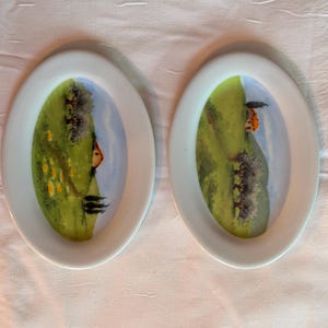 Puede incluir: Dos platos ovalados de cerámica blanca con escenas pintadas de una colina verde con una pequeña casa y árboles. Los platos están sobre una superficie blanca.
