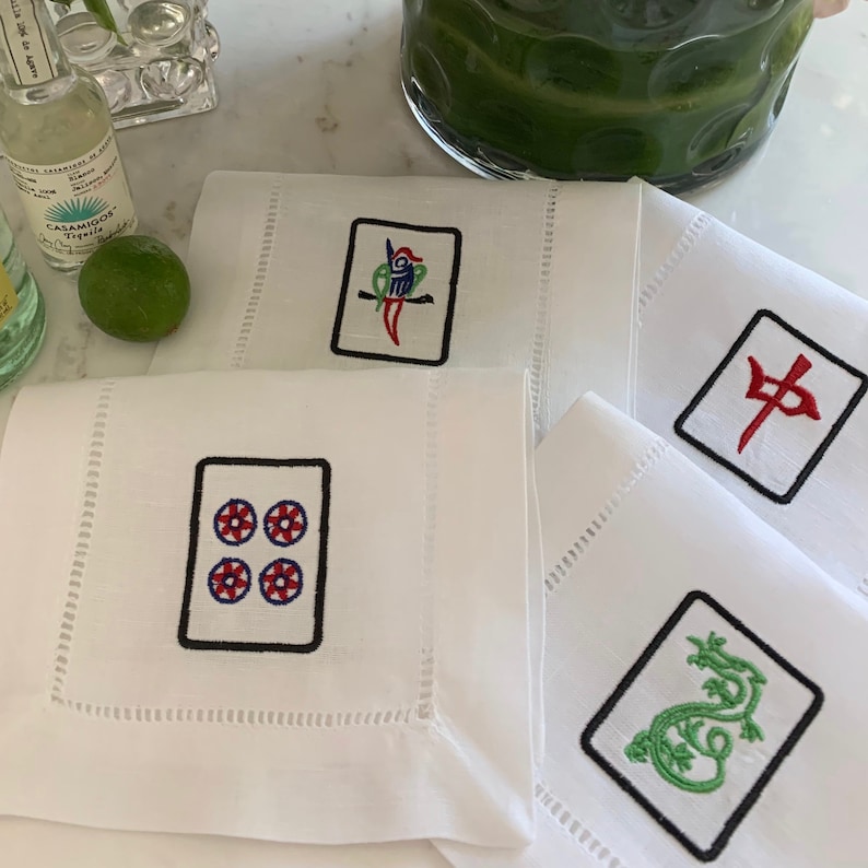 Puede incluir: Cuatro servilletas de lino blanco con fichas de mahjong chinas bordadas. Las fichas presentan un drag&oacute;n verde, un p&aacute;jaro rojo, un car&aacute;cter rojo y cuatro c&iacute;rculos rojos.