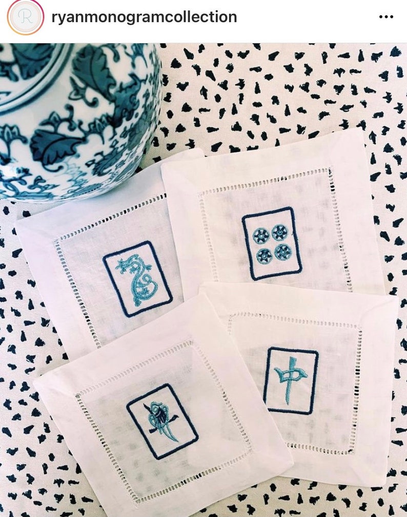 Puede incluir: Cuatro servilletas de lino blanco con dise&ntilde;os bordados en azul. Los dise&ntilde;os incluyen un drag&oacute;n, un p&aacute;jaro, un s&iacute;mbolo con una espada y un s&iacute;mbolo con cuatro c&iacute;rculos. Las servilletas est&aacute;n sobre una superficie con estampado blanco y negro.