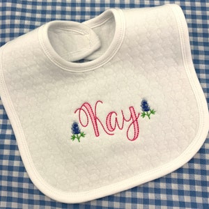 May include: White quilted baby bib with pink embroidery of the name "Kay" and blue embroidered flowers.