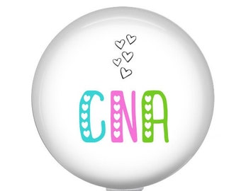 May include: A white badge reel with the letters "CNA" in teal, pink, and green, decorated with hearts. The reel has a clear plastic clip and a metal snap. The design is simple and cheerful.