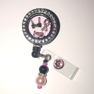 May include: A silver retractable badge holder with a pink and black breast cancer awareness charm. The charm is inside a clear round locket with rhinestones. The holder has a black beaded chain with a pink and black bead.