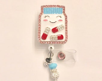 May include: A white pill bottle charm with a smiling face, pink rhinestones, and red and white pills. The charm is attached to a retractable badge holder with a blue and white beaded charm.
