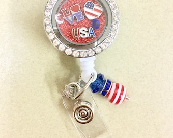 Op de afbeelding: Een zilveren intrekbare badgehouder met een transparant plastic venster. De houder heeft een ronde bedel met rode glitter en strass. De bedel heeft een rood hart met het woord "LOVE" in witte letters, een kleine Amerikaanse vlag en het woord "USA" in zilveren letters. De houder heeft ook een blauw-wit gestreepte kraal, een blauwe kristallen kraal en een zilveren bedel met een vleugelontwerp.