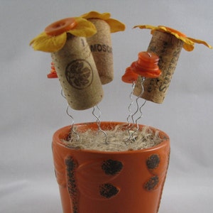 May include: A whimsical orange ceramic pot with a floral design, holding three cork flowers. Each flower features a cork body, yellow petals, and orange button accents, creating a unique decorative piece.