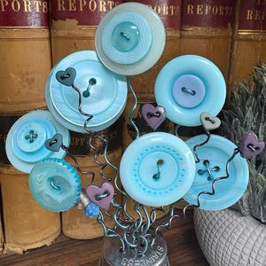 May include: A decorative arrangement of light blue buttons, some with smaller buttons in the center, attached to wire stems. Small purple heart-shaped accents are interspersed among the buttons. The arrangement is displayed in a glass jar with a stainless steel lid.