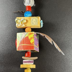 May include: A whimsical sculpture composed of various materials. It features a small, pale face at the top, followed by beads, wooden blocks, and a stone base. The sculpture incorporates a postage stamp and a butterfly embellishment.
