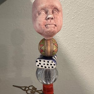 May include: A whimsical sculpture featuring a pink, expressionless face atop a stack of colorful, geometric shapes. The sculpture includes a patterned ball, a polka-dotted cube, a clear glass sphere, a red cylinder, and a blue orb. A decorative brass element extends from the side.
