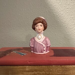 May include: A porcelain figurine of a woman's bust, featuring brown hair and a pink hat. The figurine wears a pink jacket with white buttons and a white collar. The bust is set on a red surface.