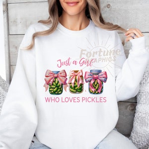 Op de afbeelding: Witte trui met de tekst "Just a Girl Who Loves Pickles" in roze letters. Drie potten met augurken met roze strikken zijn afgebeeld.