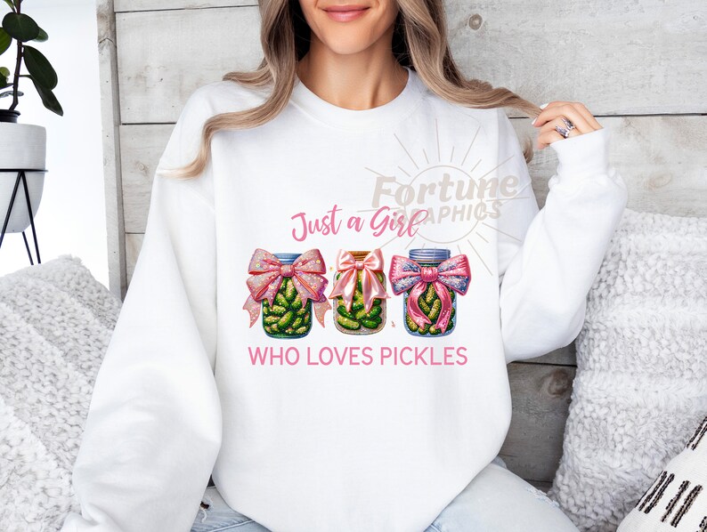 Op de afbeelding: Witte trui met de tekst "Just a Girl Who Loves Pickles" in roze letters. Drie potten met augurken met roze strikken zijn afgebeeld.