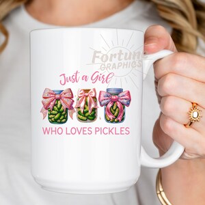 Op de afbeelding: Een witte keramische mok met een roze en groene illustratie van drie potten met augurken met roze strikken. De tekst "Just a Girl Who Loves Pickles" staat op de mok gedrukt.