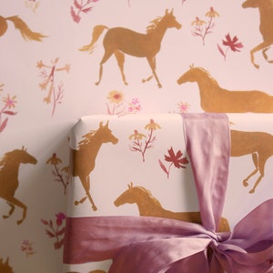 May include: A pink gift box with a pink satin ribbon tied in a bow. The box is wrapped in white paper with a repeating pattern of gold horses and pink flowers.