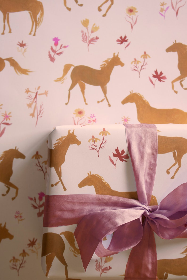 May include: A pink gift box with a pink satin ribbon tied in a bow. The box is wrapped in white paper with a repeating pattern of gold horses and pink flowers.