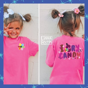 May include: A pink t-shirt with a rainbow flower on the front and the text "I CRY CANDY" with a flower, a hat, and a raccoon on the back.