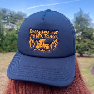 May include: A navy blue trucker hat featuring orange embroidery on the front panel. The text reads "CRASHING OUT WITH MR. TOAD" above an illustration of a character driving a vehicle flanked by flames, with "Anaheim, CA" printed below the graphic.