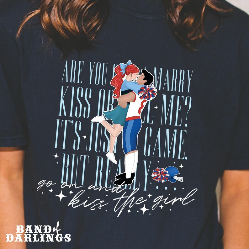 May include: A navy blue t-shirt featuring a cartoon couple kissing. The text on the shirt reads "Are you gonna marry me? It's just a game, but really go on and kiss the girl." The text "Band of Darlings" is at the bottom of the shirt.