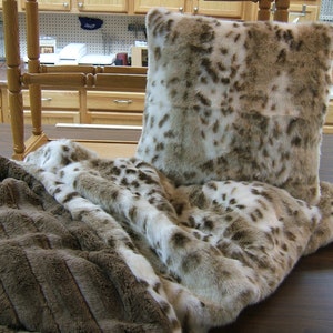 May include: A brown and white faux fur throw blanket with a matching pillow. The throw blanket is folded and draped over a wooden table.