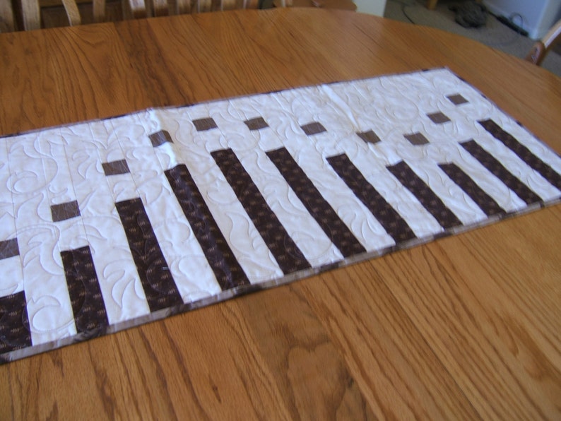 May include: A white table runner with brown stripes and squares. The runner is on a wooden table.