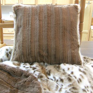 May include: A brown faux fur throw pillow with a white border. The pillow is sitting on a white and brown faux fur throw blanket.