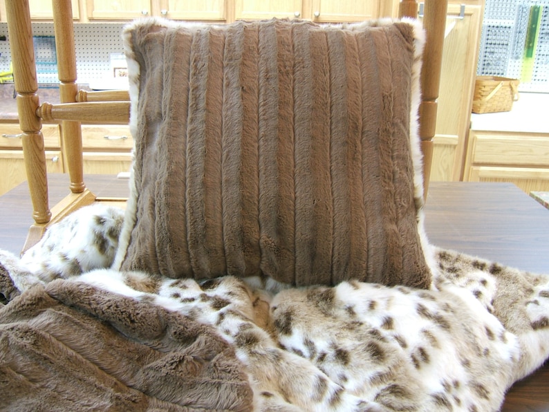 May include: A brown faux fur throw pillow with a white border. The pillow is sitting on a white and brown faux fur throw blanket.