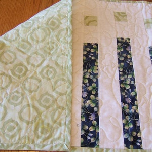 Pode incluir: Um corredor de mesa acolchoado branco com tiras de tecido floral verde e azul. O corredor de mesa tem um padr&atilde;o geom&eacute;trico verde e branco na parte de tr&aacute;s.