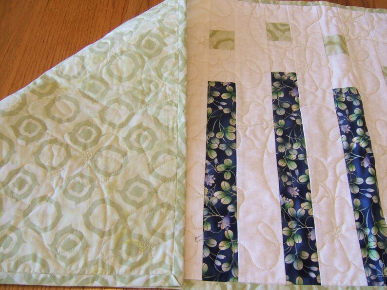 Pode incluir: Um corredor de mesa acolchoado branco com tiras de tecido floral verde e azul. O corredor de mesa tem um padr&atilde;o geom&eacute;trico verde e branco na parte de tr&aacute;s.