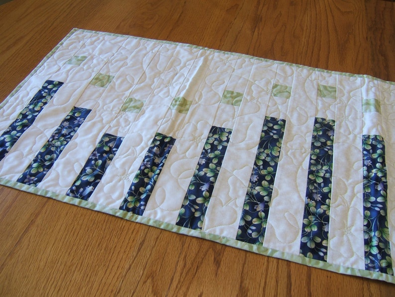 Pode incluir: Um corredor de mesa branco com uma borda verde e faixas de tecido floral azul e verde. O corredor &eacute; acolchoado com um padr&atilde;o ondulado.