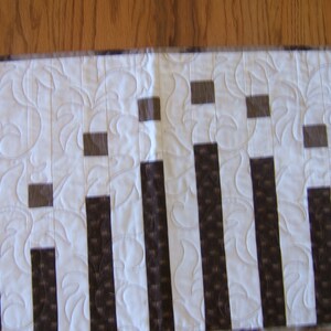 May include: A white table runner with a repeating pattern of brown vertical stripes and small brown squares. The runner has a quilted design with a floral pattern.