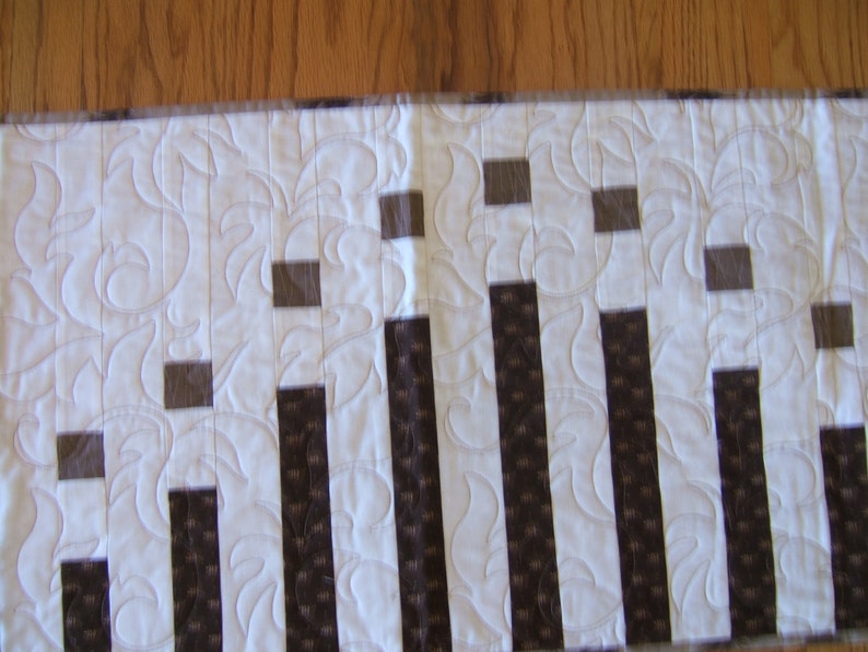 May include: A white table runner with a repeating pattern of brown vertical stripes and small brown squares. The runner has a quilted design with a floral pattern.
