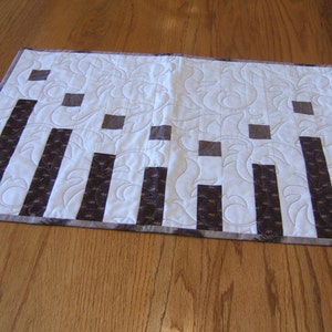 May include: A white quilted table runner with a brown and white geometric pattern. The runner has a scalloped edge and is made of fabric.