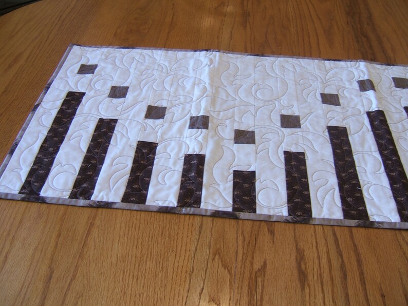 May include: A white quilted table runner with a brown and white geometric pattern. The runner has a scalloped edge and is made of fabric.